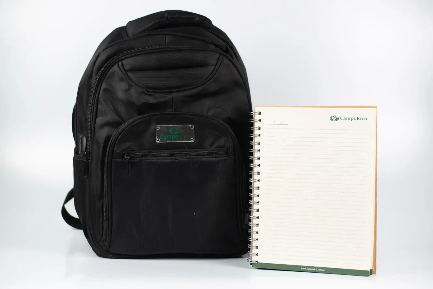 Mochila corporativa preta com placa metálica gravada, ao lado de caderno wire-o personalizado